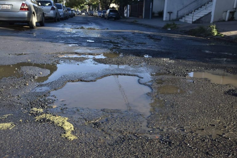 Calle de San Cristóbal con bache grande lleno de agua, pavimento deteriorado y coches estacionados en un ambiente urbano.