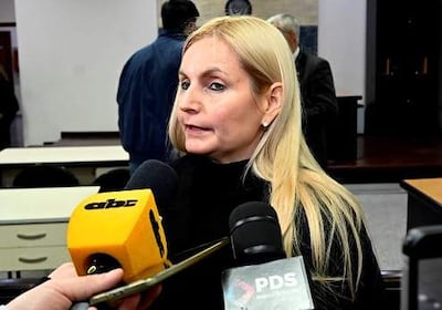 Ana Elizabeth Girala López con cabello rubio largo, en suéter negro, habla frente a varios micrófonos en una sala de conferencias.