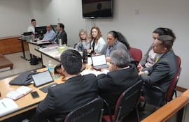 Mikhaela Rolón en blusa clara, lee una carta, mientras dos hombres con traje la observan en audiencia formal.