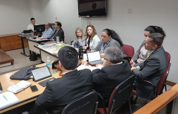 Mikhaela Rolón en blusa clara, lee una carta, mientras dos hombres con traje la observan en audiencia formal.