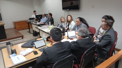 Mikhaela Rolón en blusa clara, lee una carta, mientras dos hombres con traje la observan en audiencia formal.