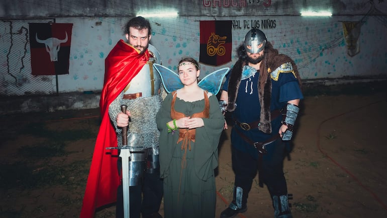 Mujer con alas de mariposa y hombres en trajes de caballero y oscuro, rodeados de decoración medieval en un evento festivo.