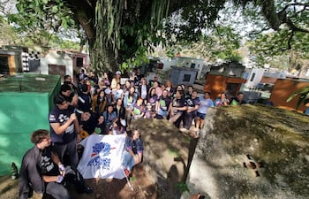 Grupo de personas con camisetas oscuras, escuchando a un hombre sentado en piedra, rodeados de árboles y tumbas en un cementerio.