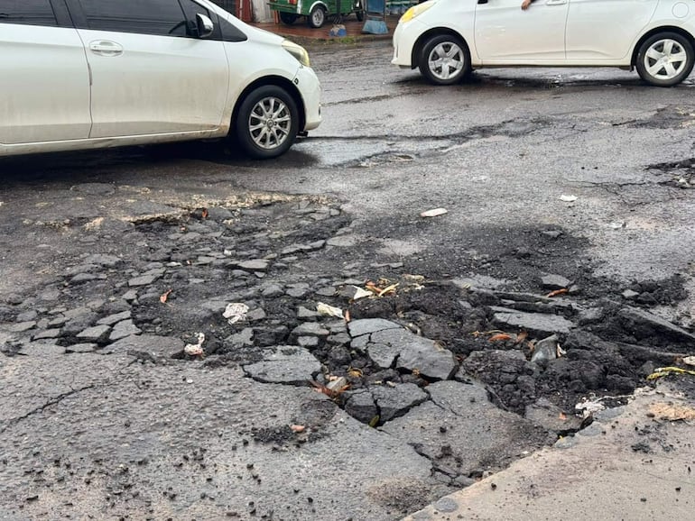 Calzada agrietada con baches, vehículos blancos estacionados y ambiente descuidado.