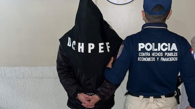 Hombre esposado de espaldas con capucha negra y letras 'DCHPEF', acompañado por agente policial en uniforme en un ambiente neutro.