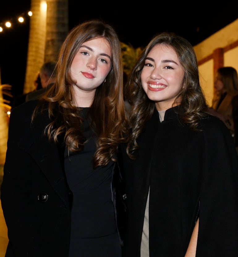 Dos mujeres sonrientes, una con chaqueta negra y otra con abrigo negro, posan en un evento nocturno con luces suaves.