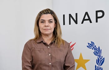 Mujer de cabello largo y rubio, vestida con camisa marrón y pantalón oscuro, frente al logo del INAP.