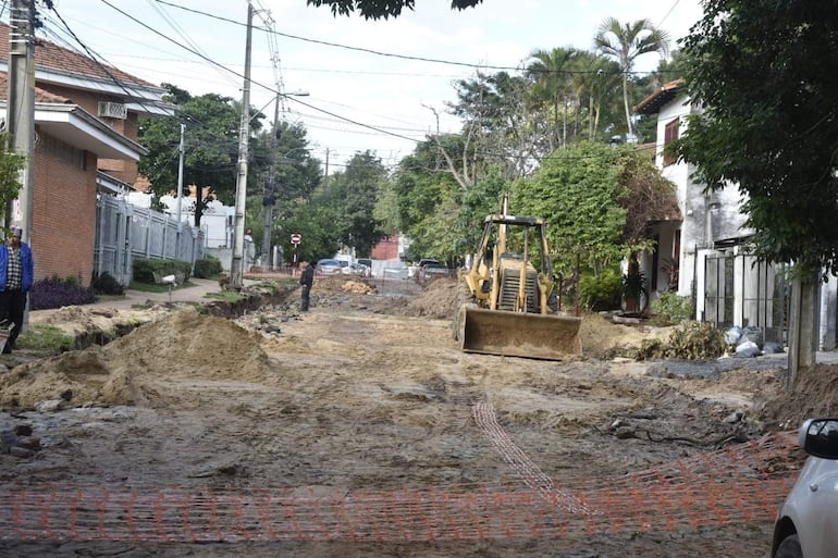 Dos trabajadores en chaleco azul y gorra, uno empujando una carretilla, con un bulldozer en una calle llena de tierra y escombros.