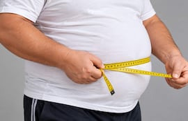 Hombre robusto con camiseta blanca y pantalones cortos mide su abdomen con una cinta métrica amarilla en fondo gris.