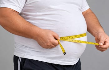 Hombre robusto con camiseta blanca y pantalones cortos mide su abdomen con una cinta métrica amarilla en fondo gris.