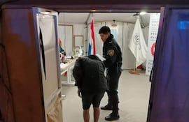 Un policía en uniforme asiste a un hombre de camiseta oscura y sandalias, en el interior de una carpa con elementos gubernamentales.