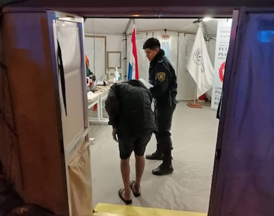 Un policía en uniforme asiste a un hombre de camiseta oscura y sandalias, en el interior de una carpa con elementos gubernamentales.