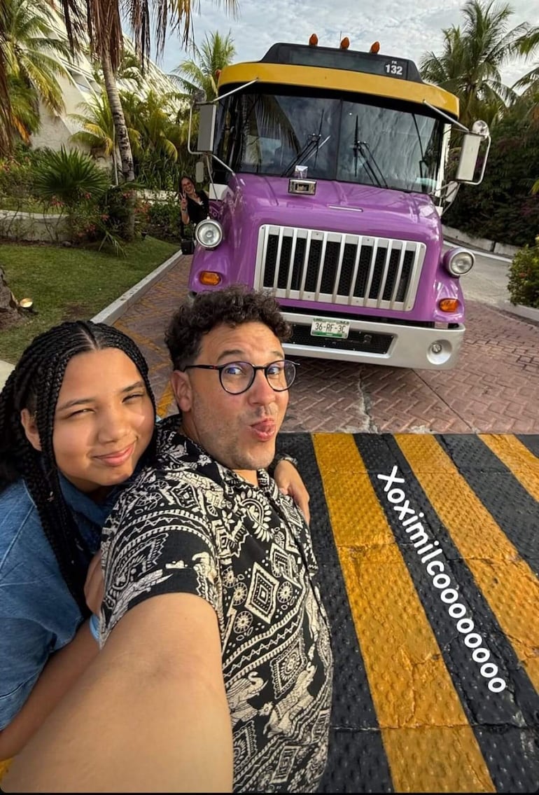 Hombre de cabello rizado con gafas y joven con trenzas en camisa azul sonríen ante un autobús púrpura en un entorno turístico.
