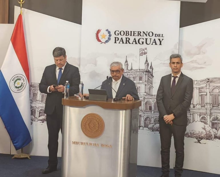 Isaías Fretes habla desde un atril, visiblemente tranquilo, mientras dos hombres lo rodean en un ambiente formal con banderas.