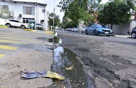 Calle con pavimento dañado y baches, vehículos estacionados y un auto azul en movimiento en un entorno urbano.