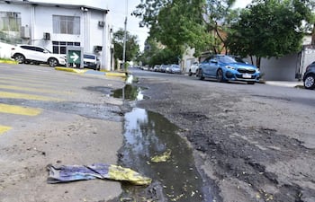 Calle con pavimento dañado y baches, vehículos estacionados y un auto azul en movimiento en un entorno urbano.