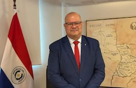 Óscar Lovera de pie en traje azul y corbata roja, junto a la bandera de Paraguay y un mapa enmarcado.