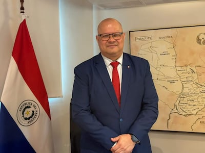 Óscar Lovera de pie en traje azul y corbata roja, junto a la bandera de Paraguay y un mapa enmarcado.