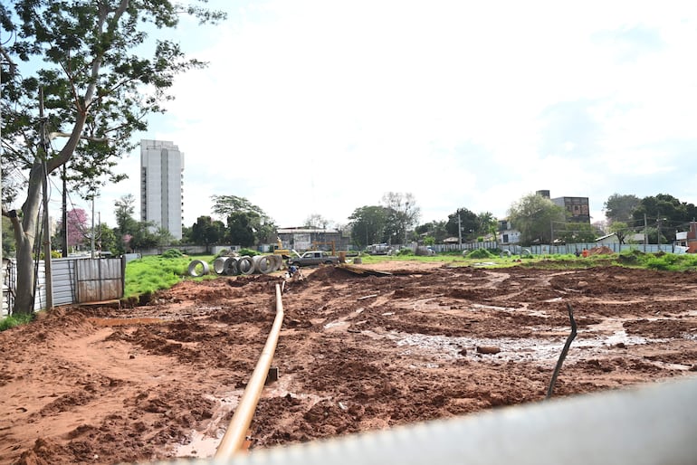 Terreno de construcción con tierra removida y maquinaria, y edificios al fondo en un entorno urbano.