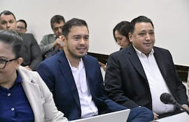 Dos hombres en trajes, uno azul y otro oscuro, sonríen mientras conversan con cuatro personas más en un ambiente serio.