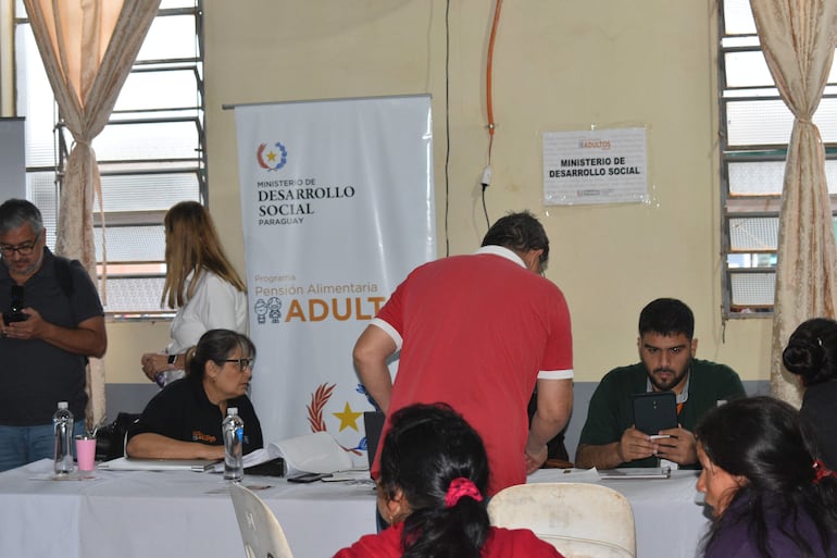 Cinco personas en espacio interior, una mujer de cabello largo atenta y un hombre en camiseta roja interactuando, documentos y botellas visibles.