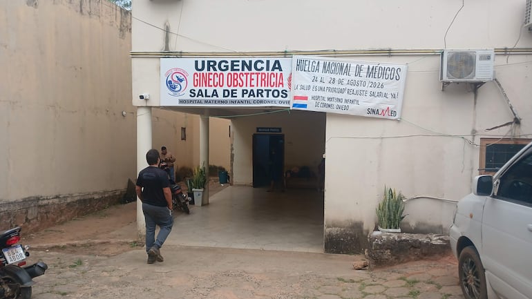 Hombres en la entrada del hospital, uno en uniforme caqui, con carteles en protesta por mejoras salariales en Neonatología.