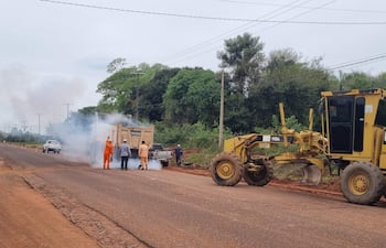 Tres trabajadores en ropa naranja y un hombre en camisa de cuadros observan un tractor CAT que opera en una carretera rural.