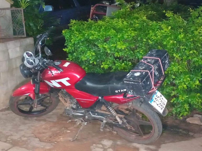 Motocicleta roja de la marca Star con cajas en la parte trasera, rodeada de vegetación en un entorno nocturno.