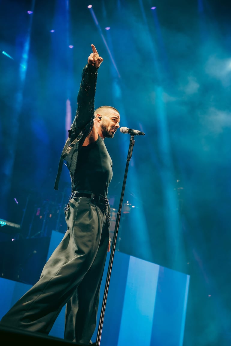 Ricky Martin de perfil en el escenario, señalando hacia arriba con un micrófono en mano, rodeado de luces brillantes.