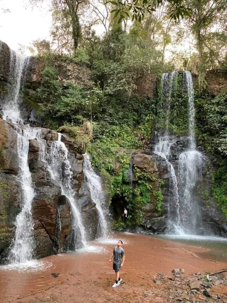 Dos personas en Salto Piraymi o Salto Jovai, ubicada en Capitán Bado; una observando la cascada desde la tierra, otra en una cueva oscura.