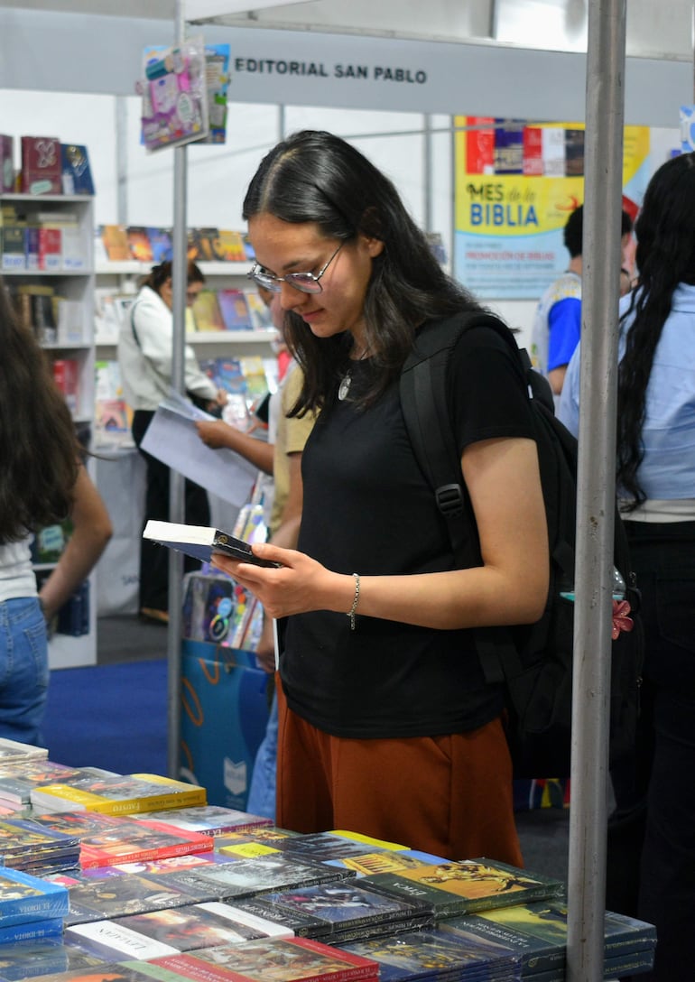 Mujer de cabello largo y oscuro, con camiseta negra y pantalones marrones, leyendo un libro en un ambiente de feria de libros.