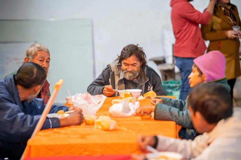 Cinco personas sentadas en mesa naranja, un hombre con barba come concentrado, una mujer joven mira al frente, ambiente cálido y social.