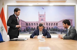 Santiago Peña firmando un decreto, vestido con traje oscuro, flanqueado por dos hombres en trajes. Ambiente de despacho gubernamental.