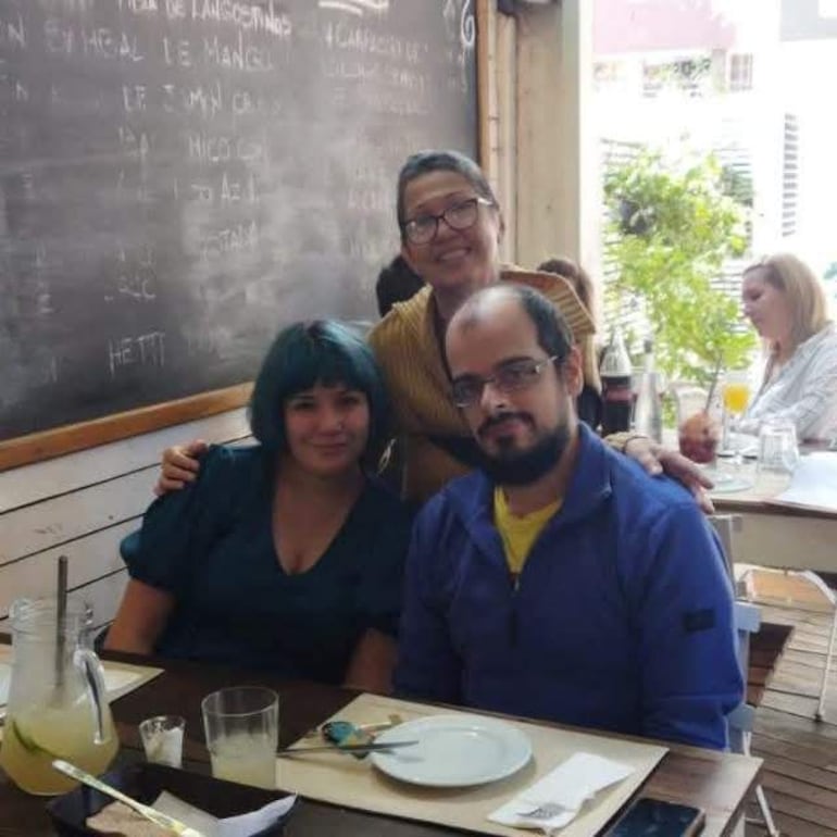Norma Beatriz con blusa oscura, sus hijas con suéter amarillo y un hombre con suéter azul, sentados en una mesa de restaurante.