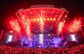 Multitud disfruta de un concierto al aire libre con fuegos artificiales, mientras músicos actúan en el escenario iluminado.