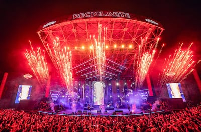 Multitud disfruta de un concierto al aire libre con fuegos artificiales, mientras músicos actúan en el escenario iluminado.