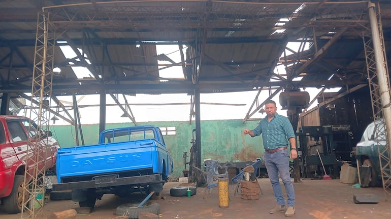 Arturo Rey Jara, vestido con camisa de cuadros, señala daños en un taller mecánico con un vehículo rojo y camioneta Datsun al fondo.