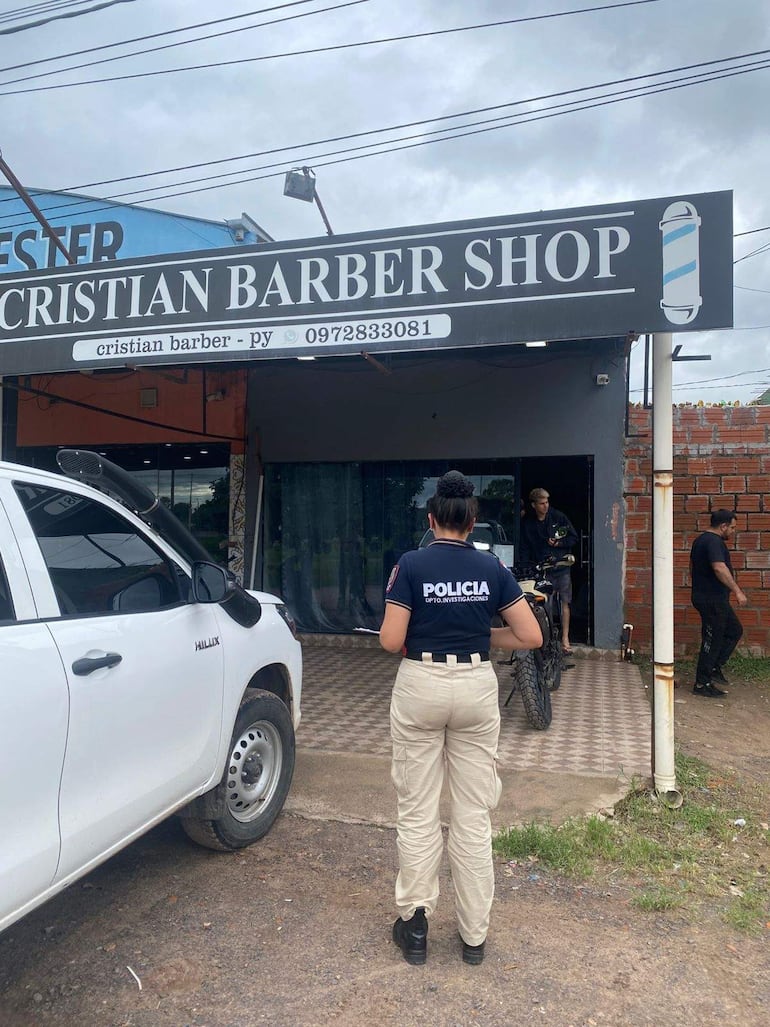 Mujer policía con camiseta POLICÍA se acerca al barber shop, mientras un vehículo policial está estacionado a su izquierda.
