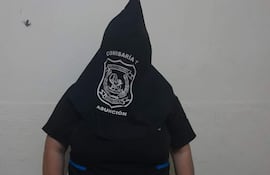 Persona de pie con capucha negra y camiseta con logotipo de la policía en fondo blanco, sin otros individuos visibles.