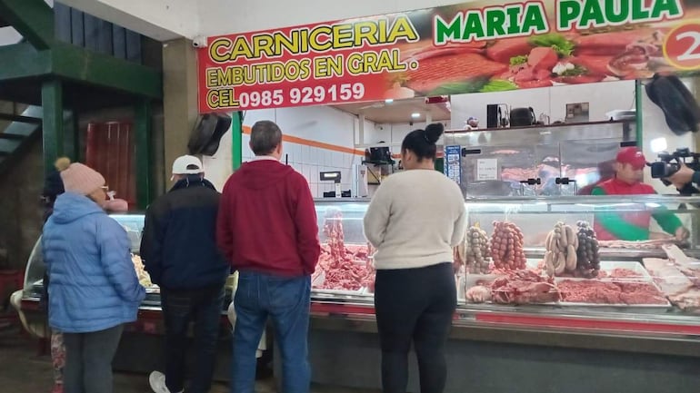 Clientes en carnicería María Paula, algunos con gorro rosa y abrigo azul, observan embutidos tras el mostrador de un carnicero vestido de rojo y verde.