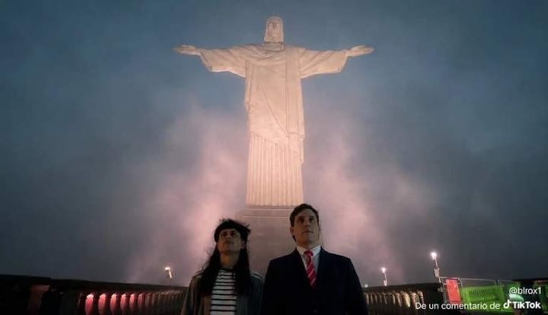 Gaspi en traje oscuro y una mujer con blusa a rayas y chaqueta negra, contemplan la iluminada estatua del Cristo Redentor en una noche con niebla.