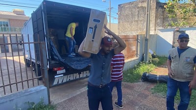 Hombres en camisetas de diversos colores descargan urnas electrónicas del camión, en un ambiente exterior tranquilo.