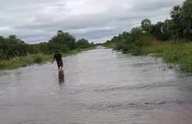 Persona con vestimenta oscura camina descalza por un camino inundado, rodeada de vegetación y cielo nublado.