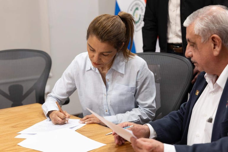Rossana Gonzáñez con camisa clara firmando documentos, mientras Isaías Fretes con papeles observa atentamente en fondo con banderas.