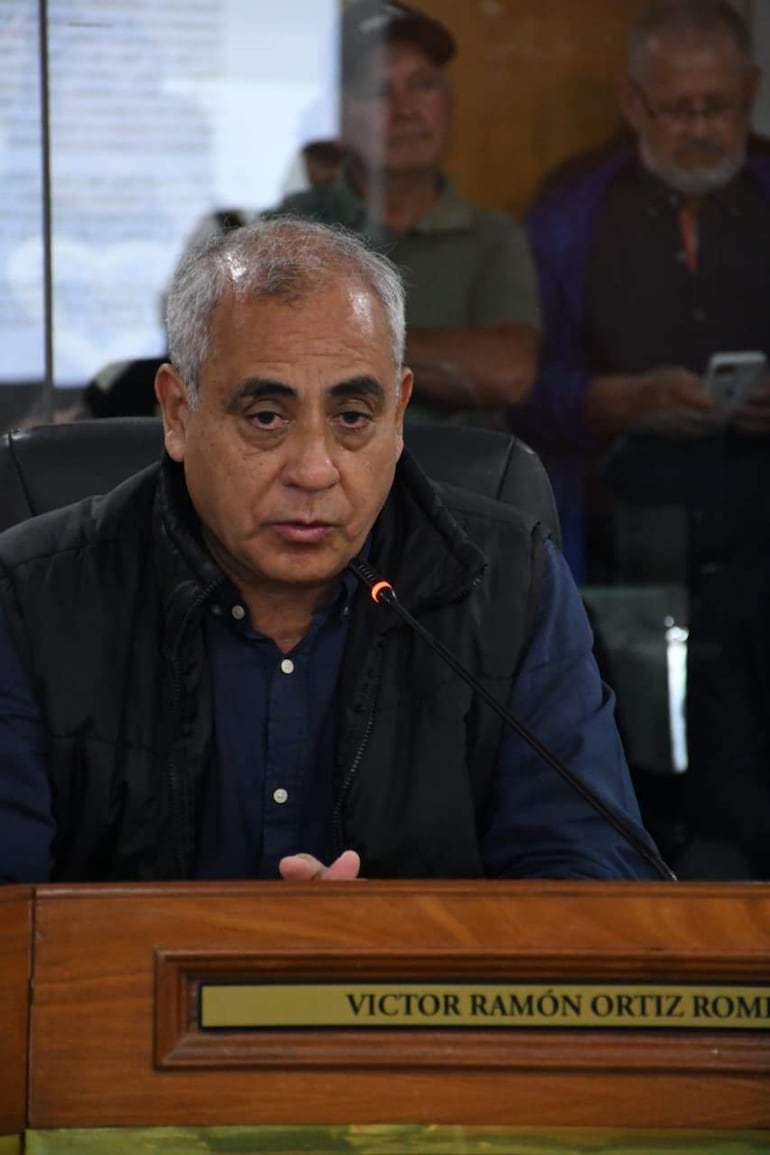 Víctor Ramón Ortiz Rom, con chaleco negro y camisa azul, expone ante el micrófono en una sala, rodeado de público atento.