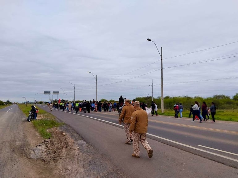 Manifestantes vestidos de varios colores marchan en carretera con banderas, acompañados por dos autoridades de seguridad en uniforme.