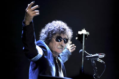 Charly García con cabello rizado y barba, usando gafas de sol y chaqueta oscura, levantando los brazos en un escenario iluminado.