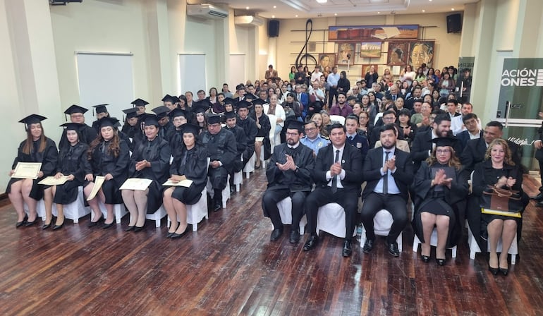 Cuarenta graduados con togas negras en un salón, algunos sosteniendo diplomas, mientras sus familiares aplauden con sonrisas.