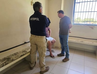 Agente de INTERPOL con chaqueta negra de espaldas, conversa un hombre en camiseta color oliva y otro sentado en camiseta púrpura.