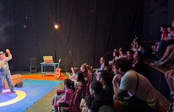 Hombre de mediana edad, vestido de overol y camisa de cuadros, actúa en el escenario, rodeado de un público atento y animado.
